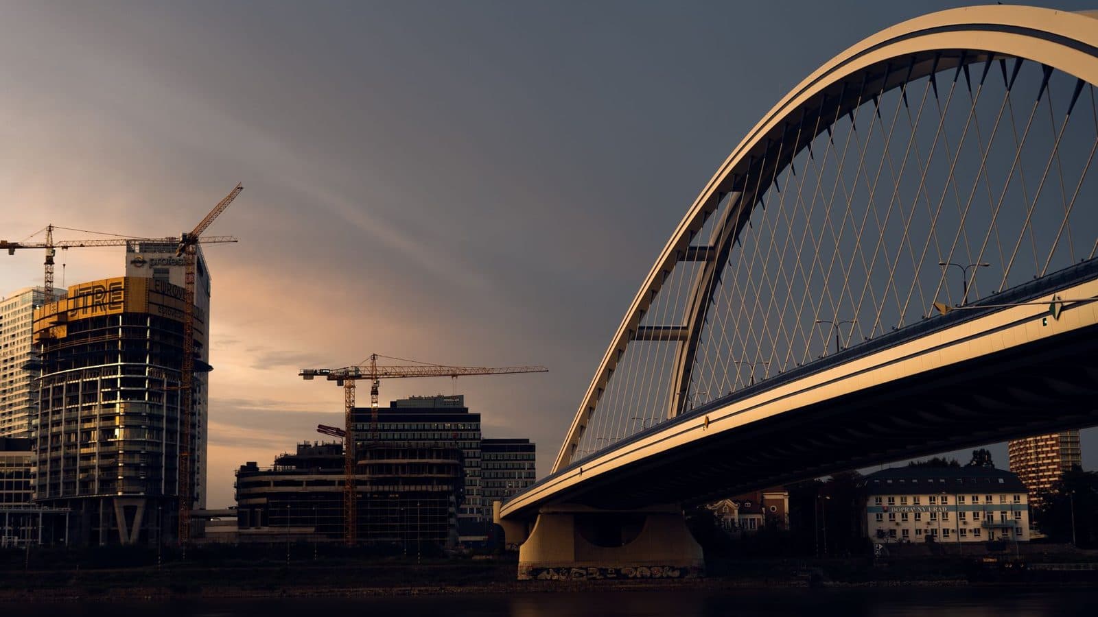 Ponte urbana em construção ao entardecer, com guindastes ao fundo