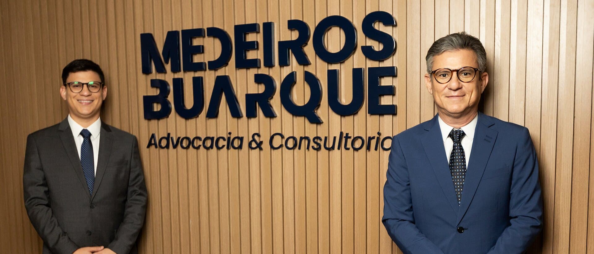 Recepção da sede, no Centenário Office: o letreiro Medeiros Buarque Advocacia & Consultoria no painel ripado de madeira, com os sócios Caio Lucas Buarque e Dalton Medeiros Buarque um de cada lado.